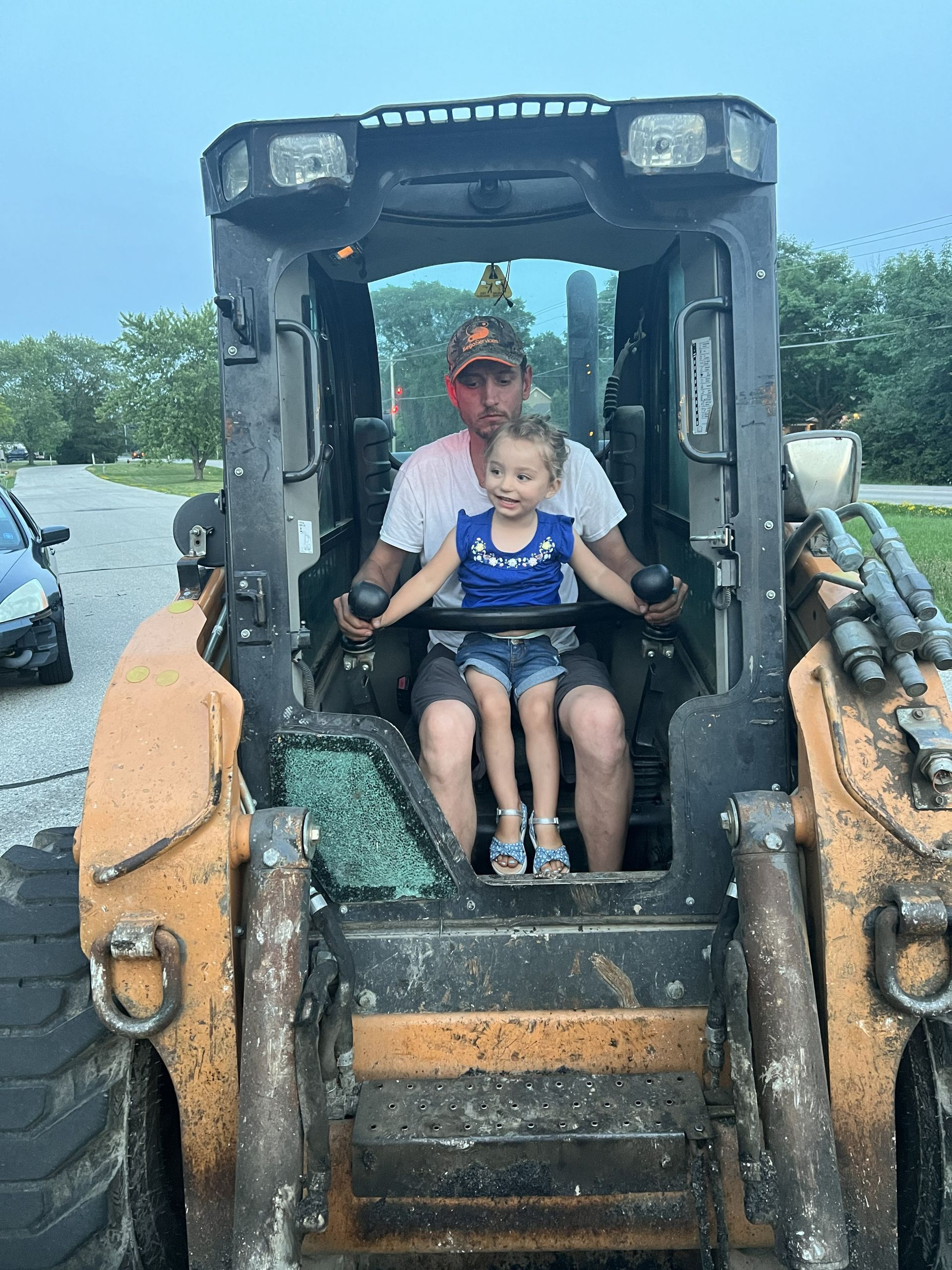 Child sitting on a muddy skid steer with an adult operator in the cab outdoors