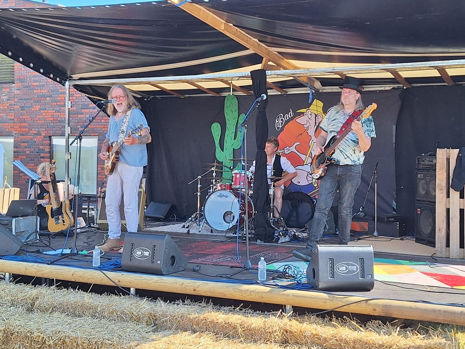 Band treedt op een openluchtpodium op: gitaristen, drummer. Cactussen op de achtergrond, zwart bladerdak, zonnige dag.
