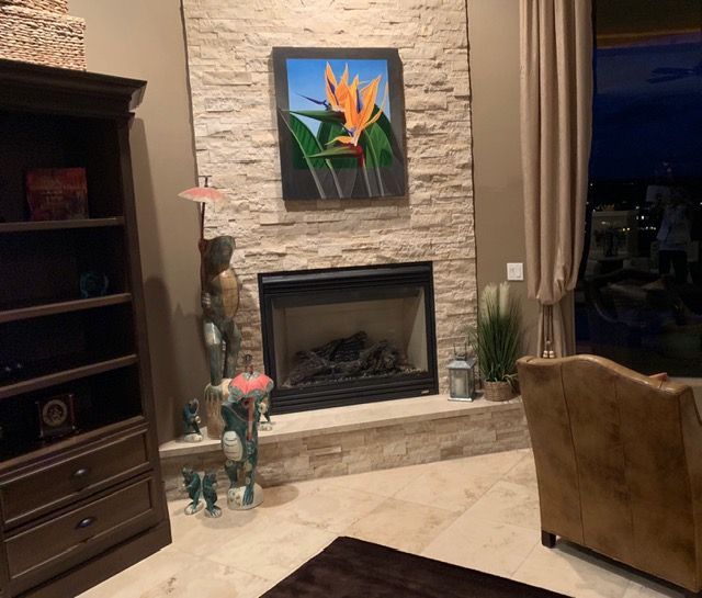 Living room with stone fireplace, bird painting, beige curtains, and a brown chair at right