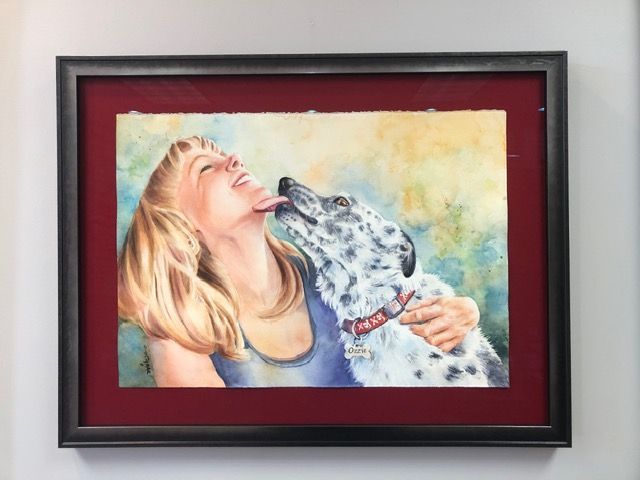 Framed painting of a blonde woman smiling as she kisses a spotted dog, with a red mat and dark frame