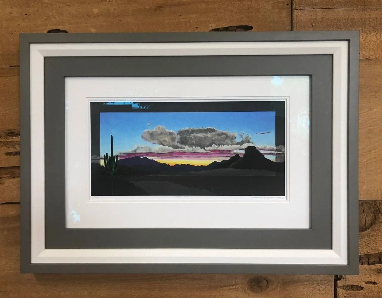 Framed sunset landscape print with mountains and colorful sky on a wooden wall.