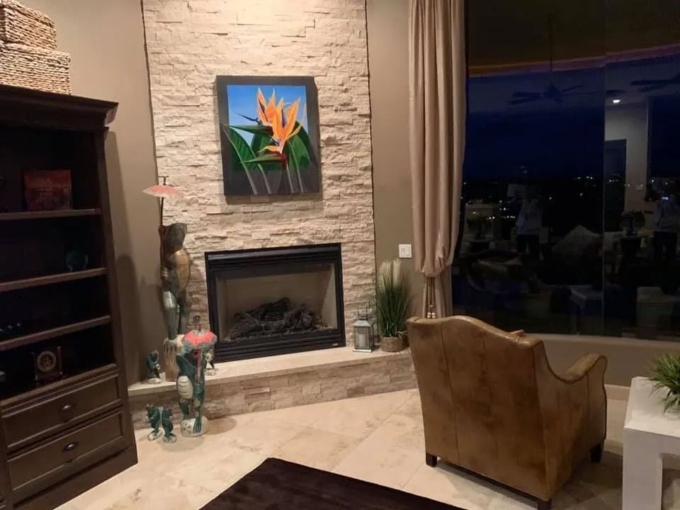 Living room with stone fireplace, floral painting, dark bookshelf, and tan chair overlooking a night view.