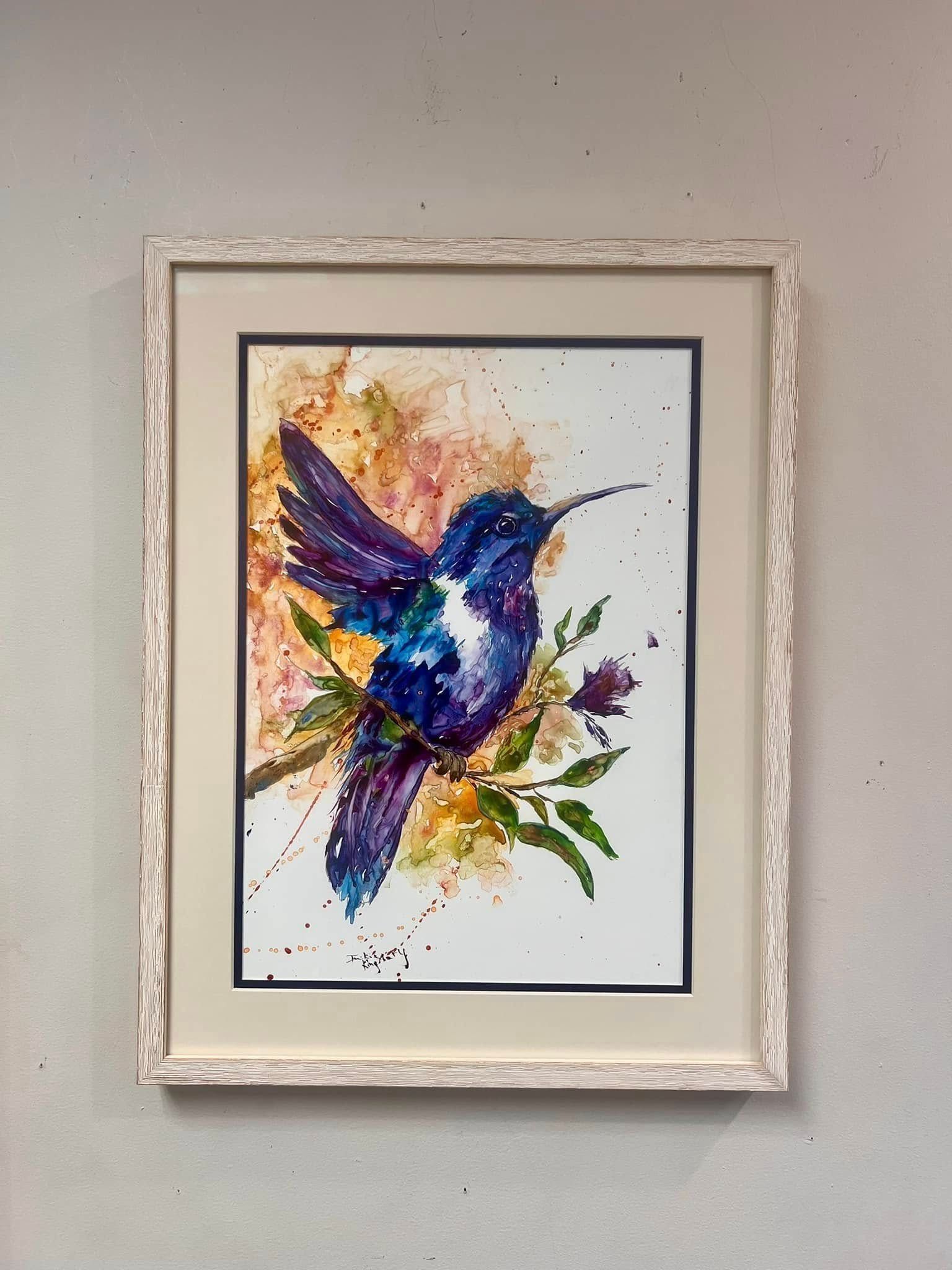 Framed watercolor hummingbird painting with purple, blue, and green splashes on a white wall