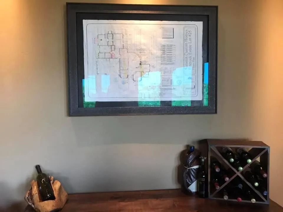 Framed print on a gray wall above a wooden desk with a wine rack and decorative bottles below