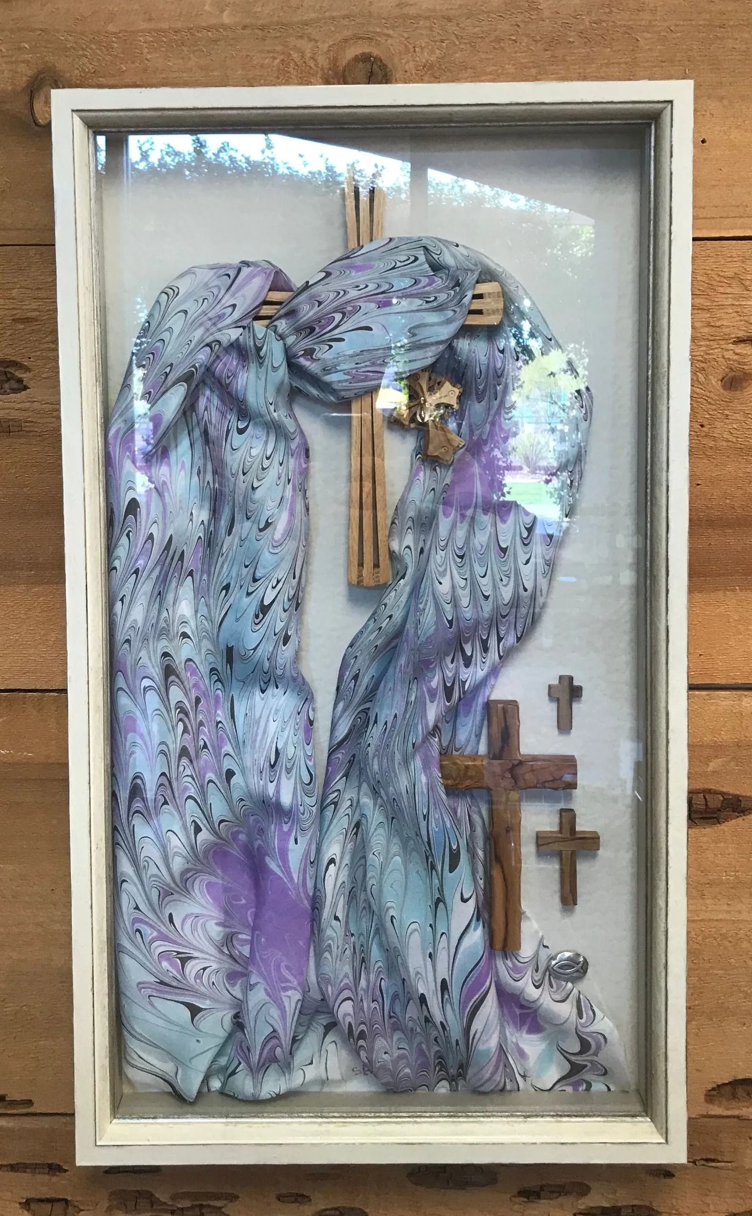 Purple angel statue with wings in a shadow box, beside three small crosses and a window reflection