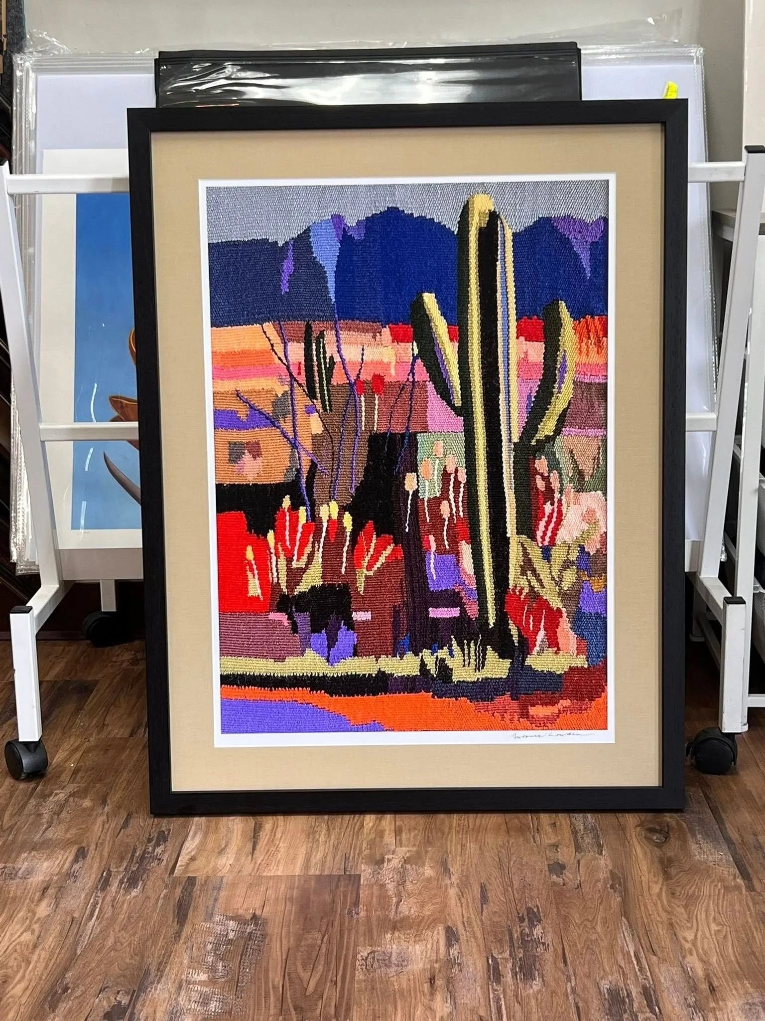 Framed colorful desert cactus artwork with bold reds, yellows, and blue sky on a wood floor.