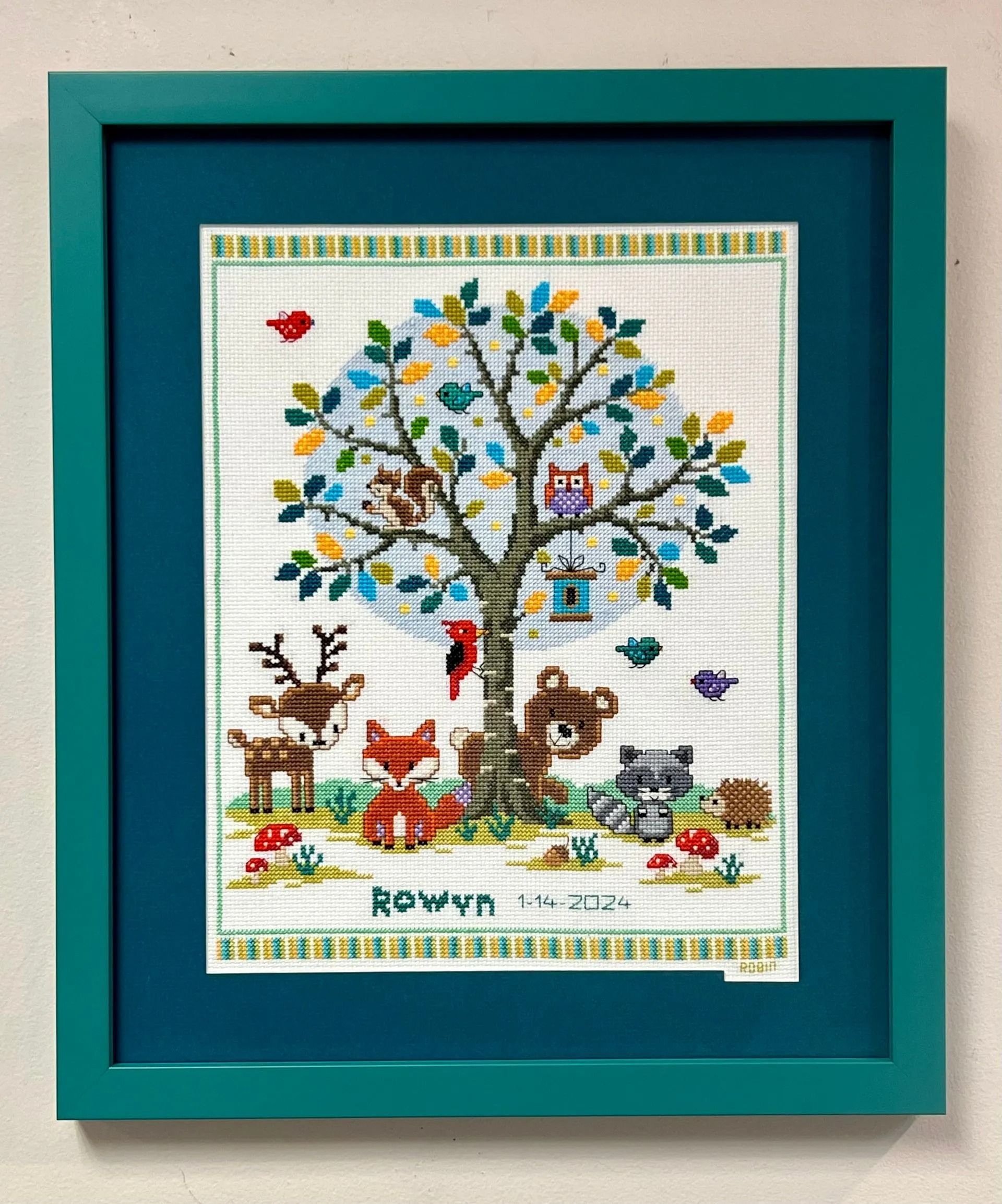 Framed embroidered woodland scene with animals around a tree on a teal mat.