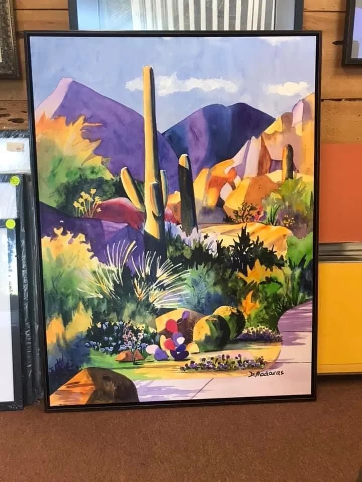 Colorful desert landscape painting with purple mountains, cactus, flowers, and a bright sky in a black frame