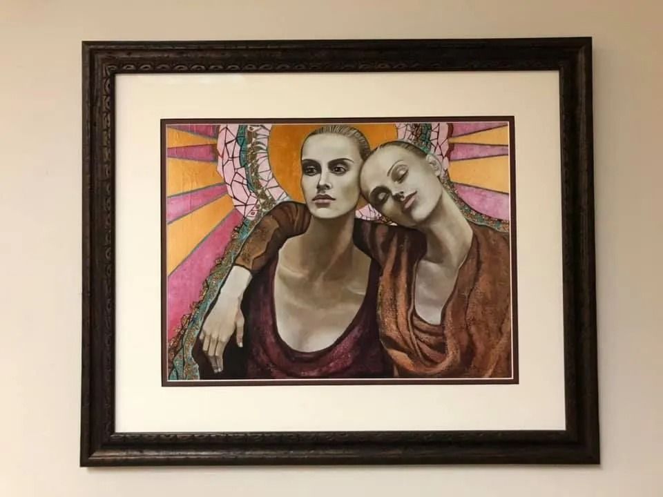 Framed artwork of two stylized figures, one leaning on the other, with pink and orange sunburst background
