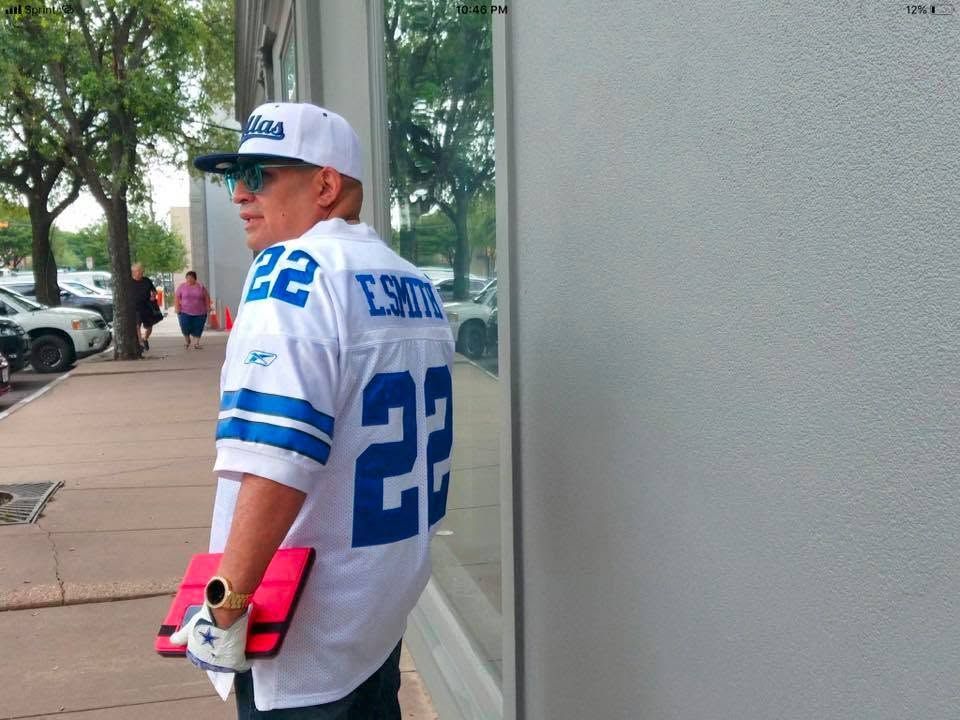 Person in a white and blue jersey and cap, standing on a sidewalk beside a building and looking back.