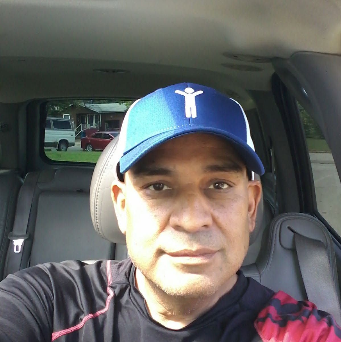 Man sitting in a car, wearing a blue cap and dark shirt, looking at the camera.