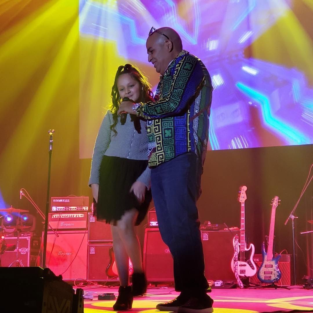 Two performers dancing onstage under colorful concert lights, with guitars and drums in the background