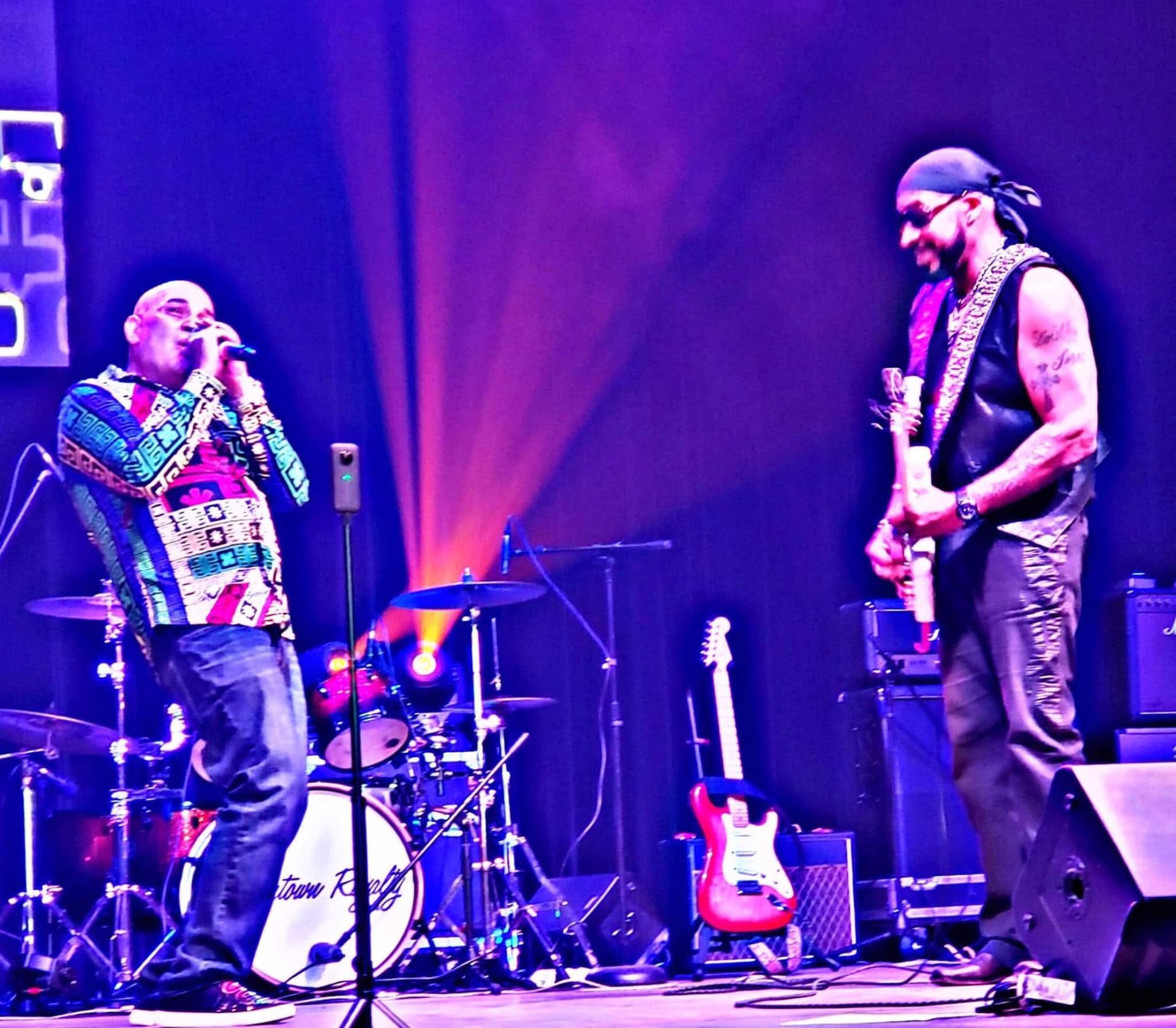 Two musicians performing onstage under purple lighting, with drums and guitars in the background.