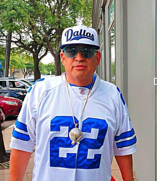 Person in a white Dallas #22 jersey and cap, wearing sunglasses, standing outdoors by a sidewalk.