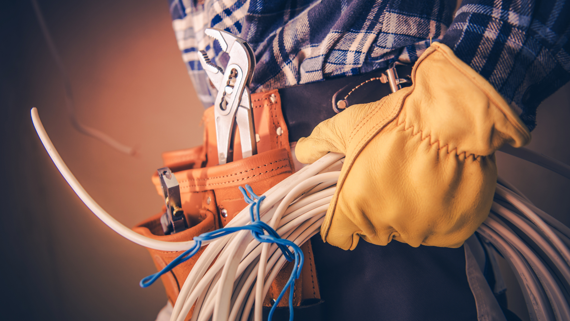 A person wearing a plaid shirt and tool belt holds a coil of white electrical wire while wearing a yellow work glove.