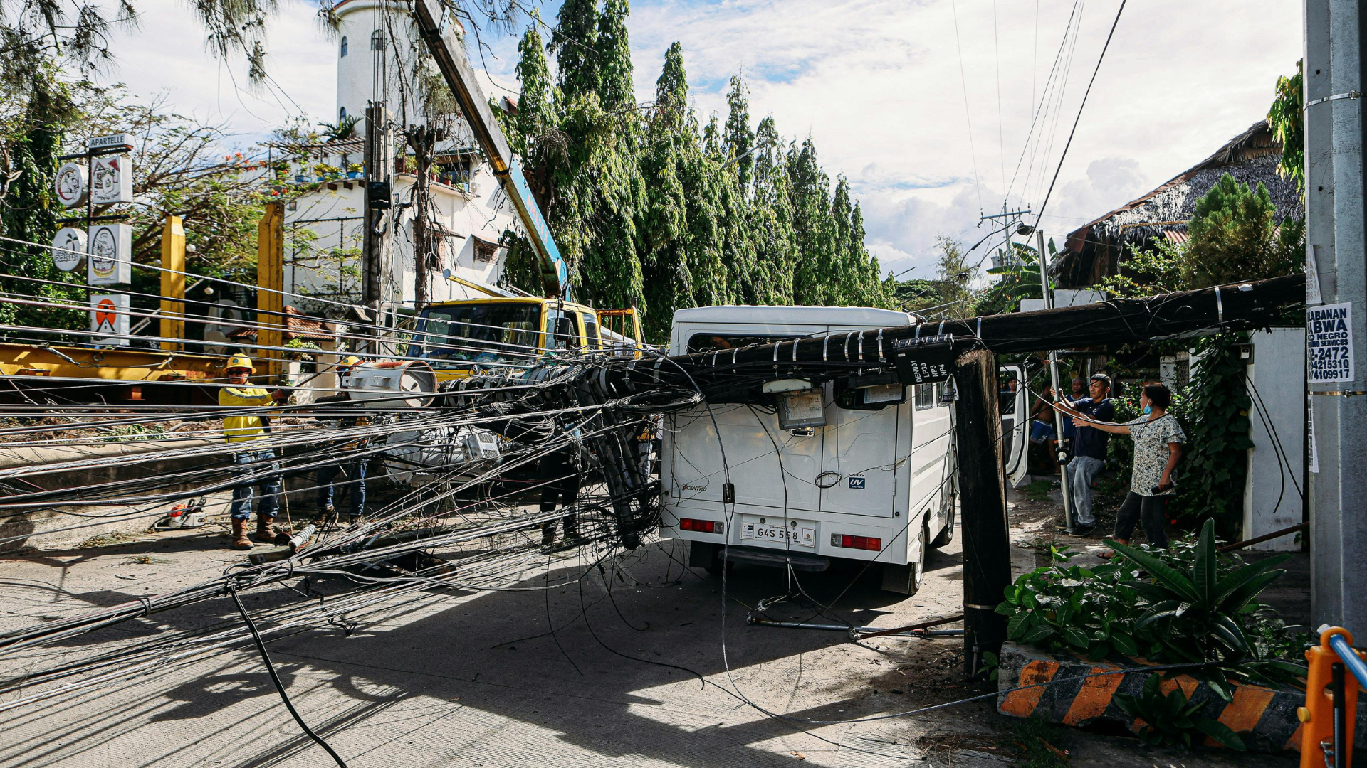 A crane boom has collapsed onto a white van parked outdoors, with a worker standing nearby in a construction area.