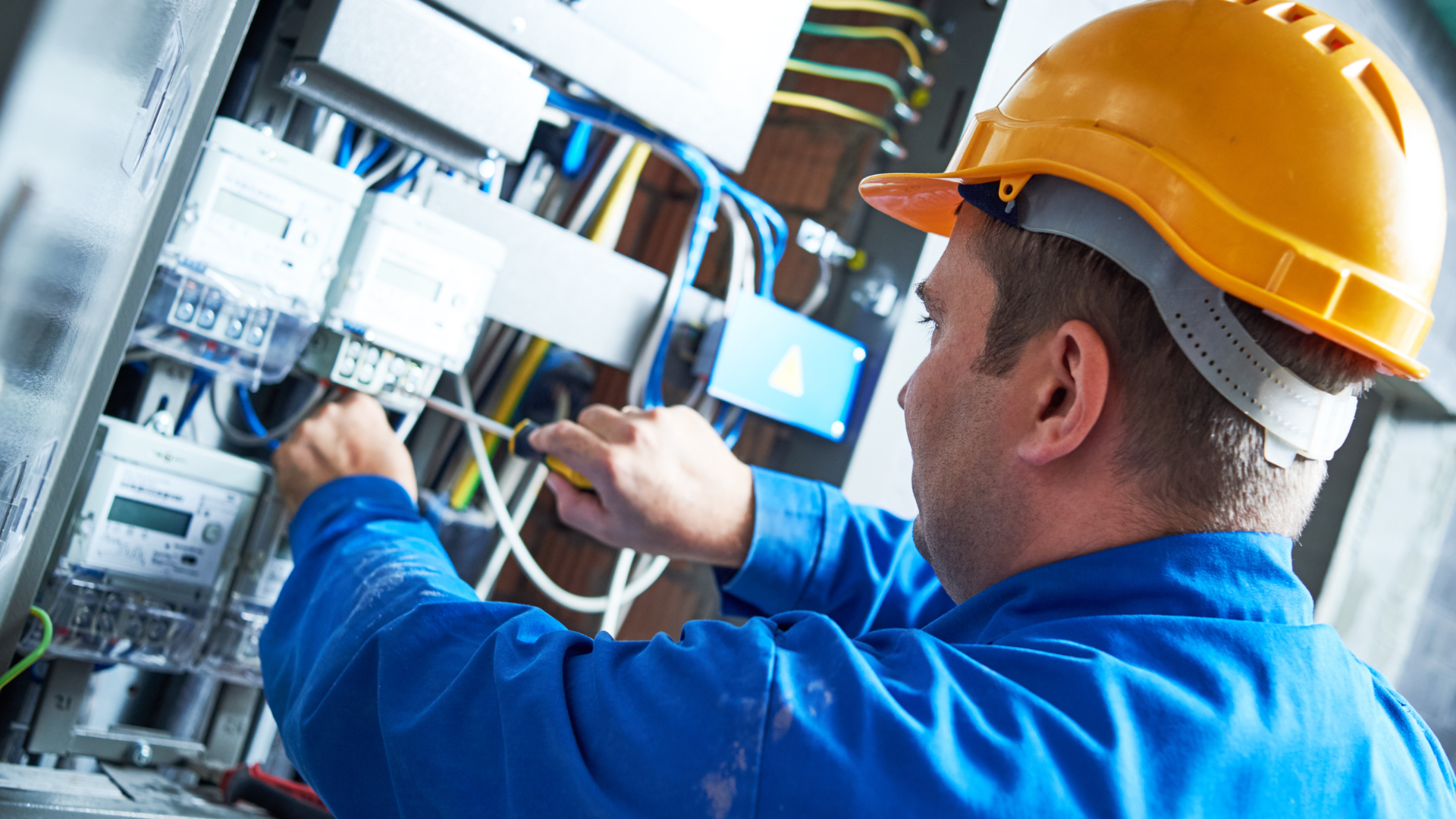 A technician in a yellow hard hat and blue uniform uses a screwdriver to work on electrical wiring in a utility box.