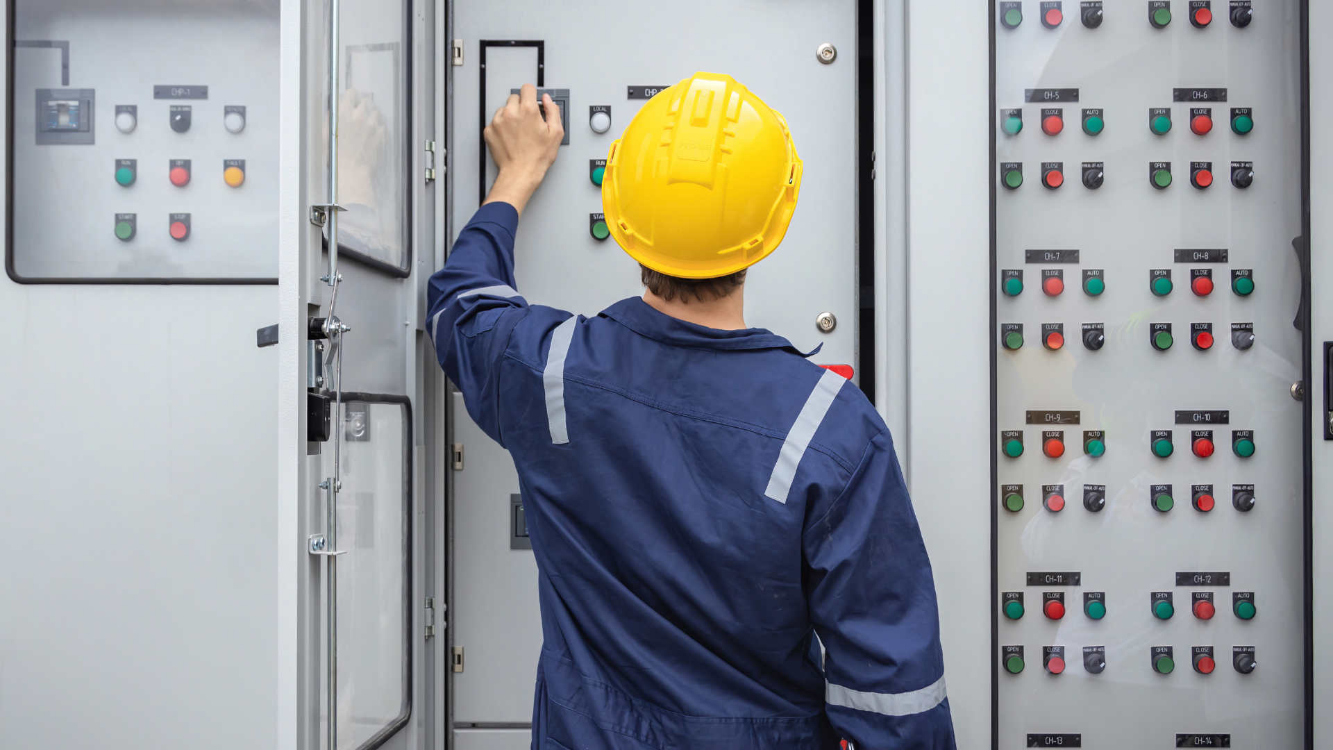 A worker in a blue uniform and yellow hard hat adjusting controls on a large industrial electrical panel.
