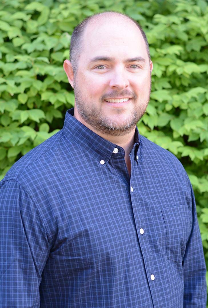 A man in a blue shirt is smiling in front of a bush.