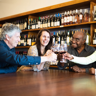 Friends clink wine glasses at a cozy bar, smiling across a wooden table.