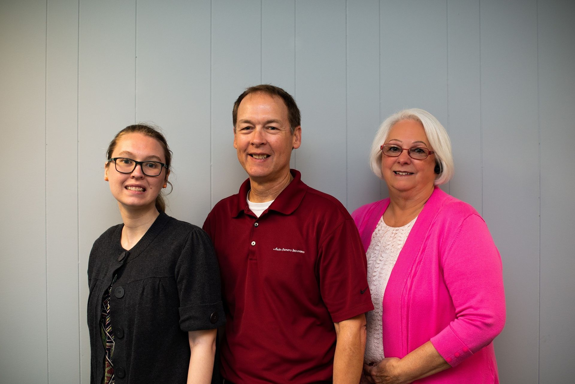Three people smiling in front of a gray wall: a young woman in glasses, a man in red, and an older woman in pink.