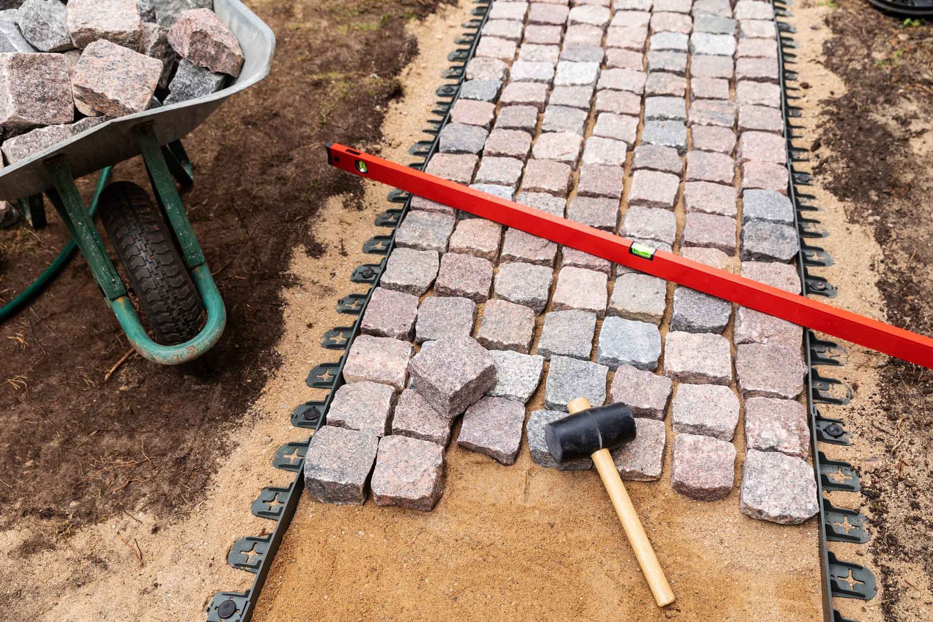 Paver path under construction with stones, red level, and hammer beside a wheelbarrow