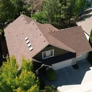 Aerial view of a suburban house with brown roof and dark siding, surrounded by trees and a driveway.