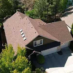Aerial view of a suburban house with brown roof and dark siding, surrounded by trees and a driveway.
