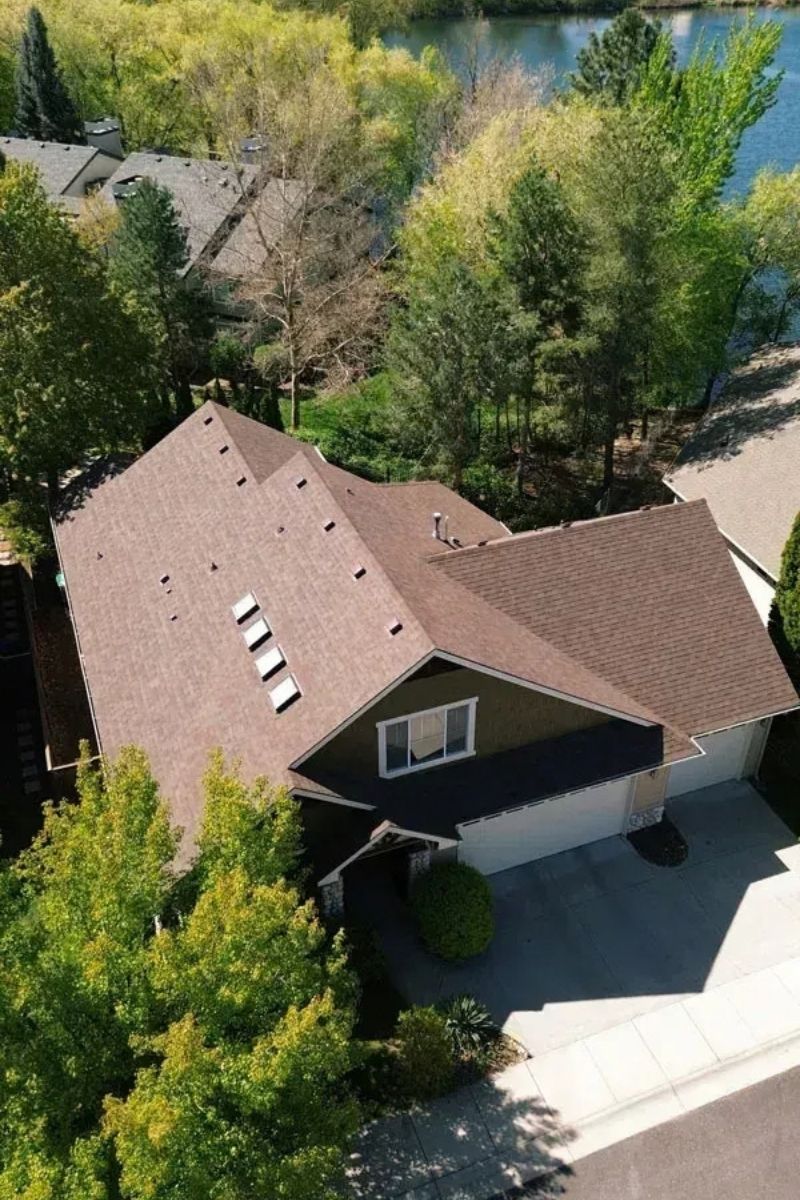 Aerial view of a brown-roofed house surrounded by green trees near a lake.