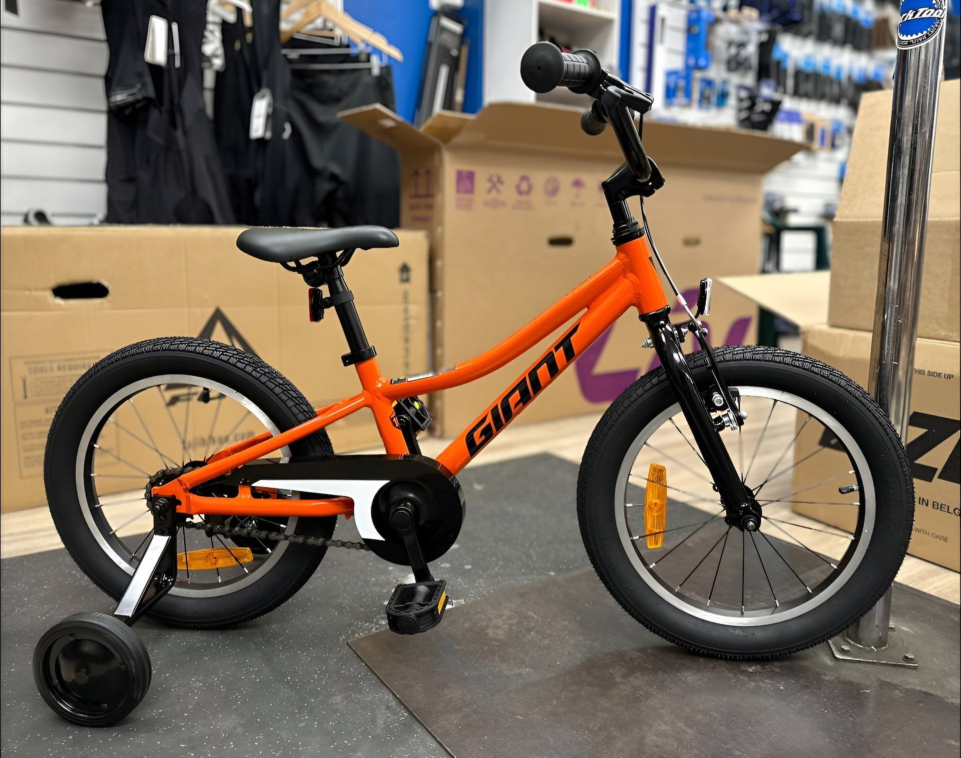 Orange Giant kids' bicycle with training wheels, black tires, and black handlebars, in a shop — Grafton City Cycles in Grafton, NSW