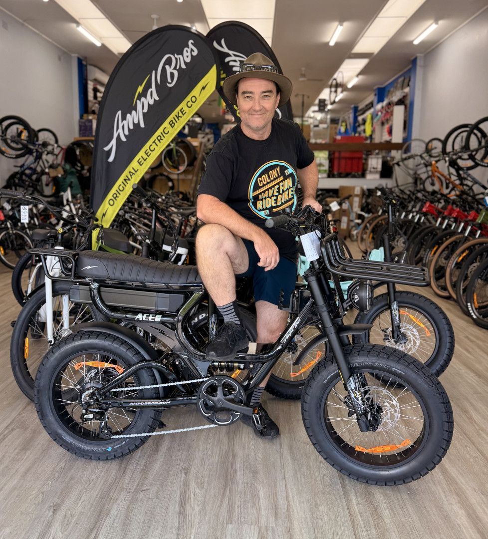 Man seated on black electric bike inside a bike shop, leaning on handlebars, wearing a hat — Grafton City Cycles in Grafton, NSW