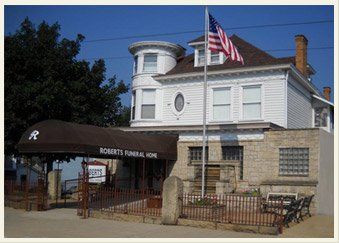 Roberts Funeral Home building with white siding, black awning, and American flag on a sunny day