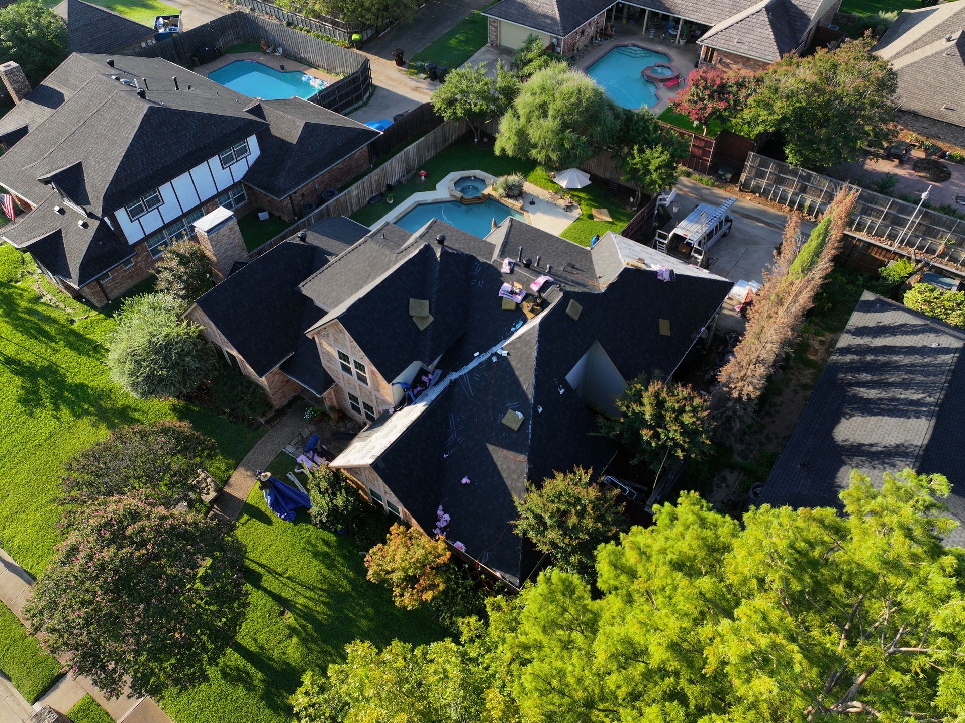 Aerial view of houses with dark roofs, pools, and green lawns in a suburban neighborhood.