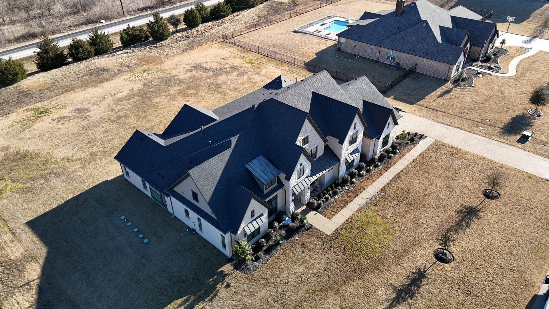 Divine Construct | Aerial view of a large, dark-roofed house with a long driveway and surrounding dry grass.