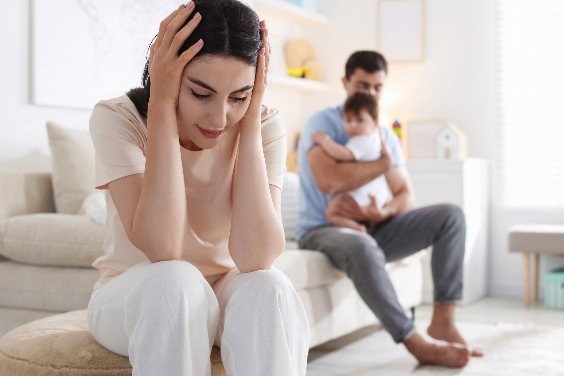 Woman sits upset in foreground while man holds baby in background, suggesting family conflict