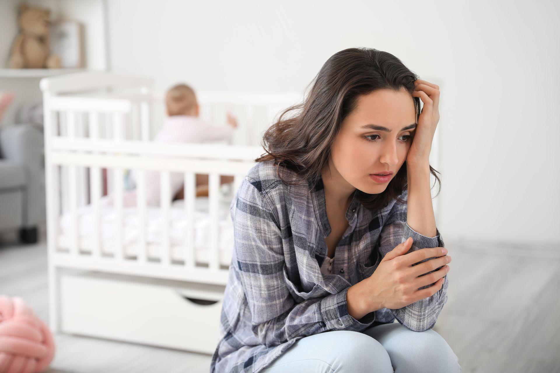 Woman sitting on the floor by a crib, looking distressed and holding her arm in a bright nursery.