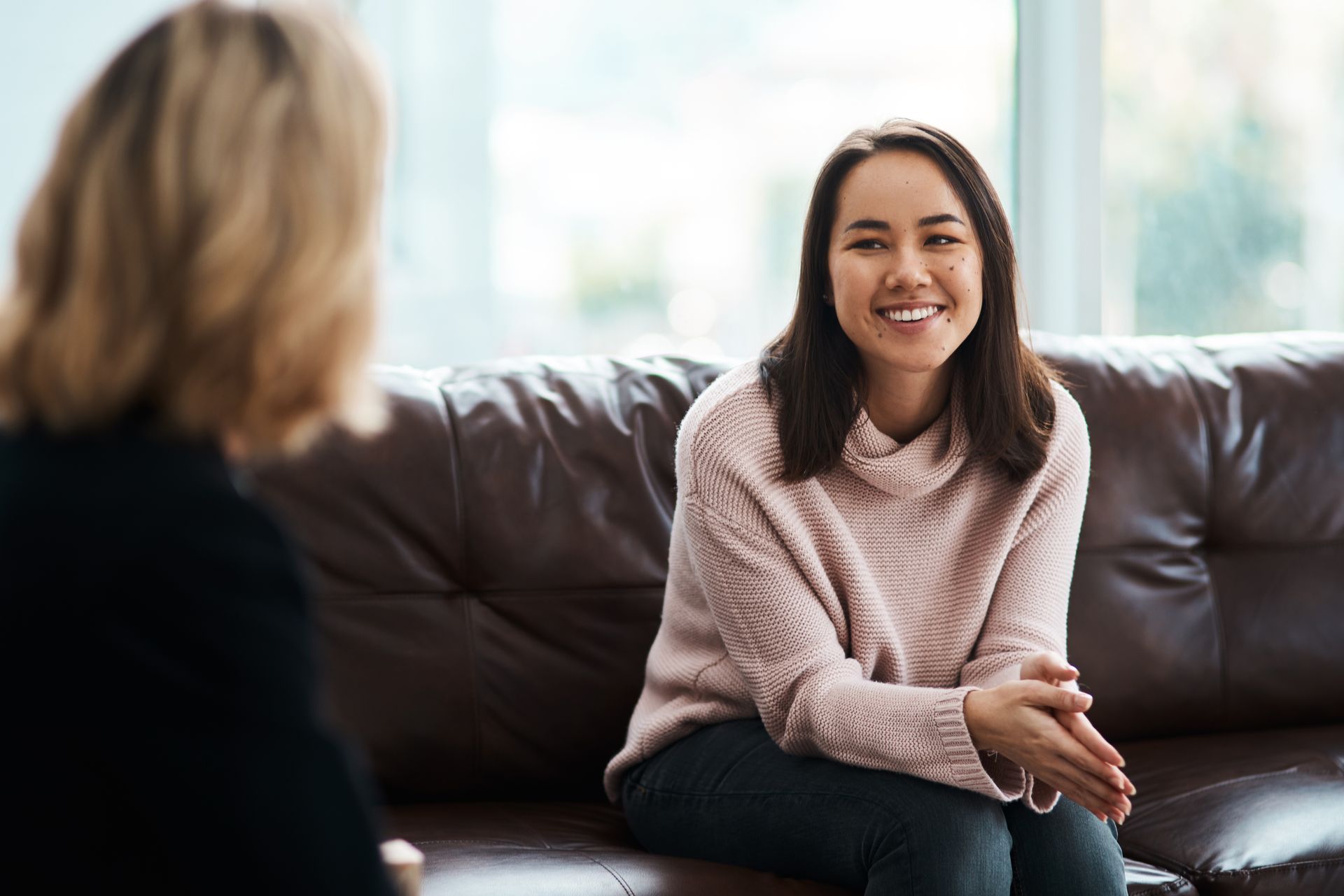 Two women talking on a couch, one smiling in a pink sweater in a bright room.