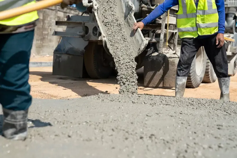 Construction workers pouring wet concrete on a road beside heavy machinery, wearing reflective safety vests.