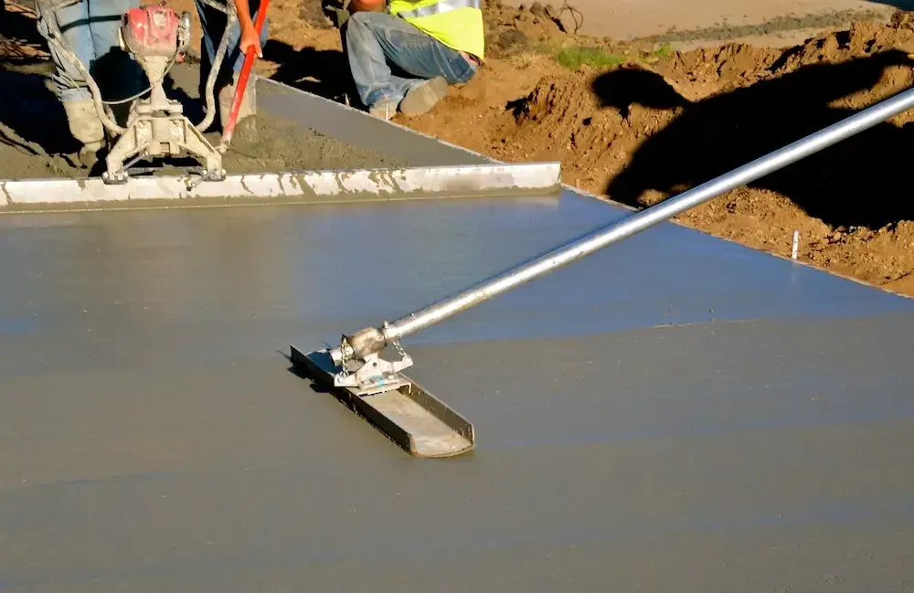 Worker smoothing fresh concrete with a long-handled screed at a construction site