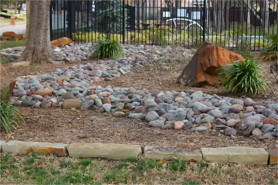 Rock garden with smooth stones, tree stump, and ornamental grasses beside a black fence