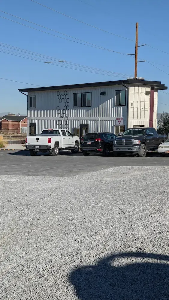 Small white building with striped siding, parked cars, and a utility pole on a gravel lot under a clear sky