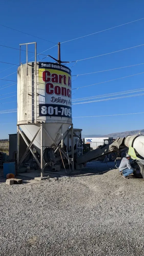 Industrial gravel yard with a white silo, signage, and concrete mixer trucks under a clear blue sky.