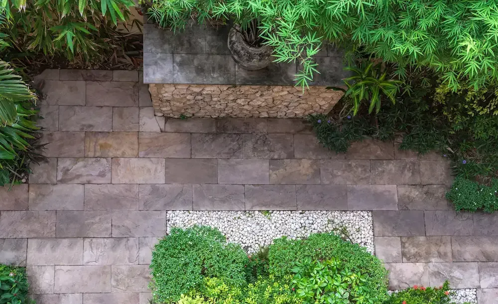Stone patio with a small central fountain, bordered by green shrubs and trees