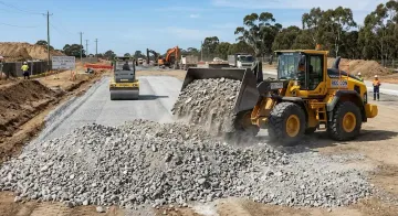Road construction with a bulldozer dumping gravel onto a new roadbed, with a roller in the background.