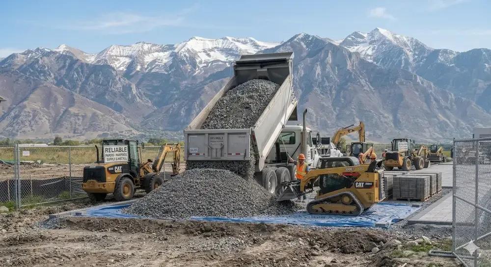 Bulldozers and a dump truck unload gravel at a construction site with snow-capped mountains behind.