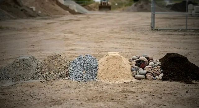 Two large gravel piles, one tan and one gray, in a gravel yard with trees in the background