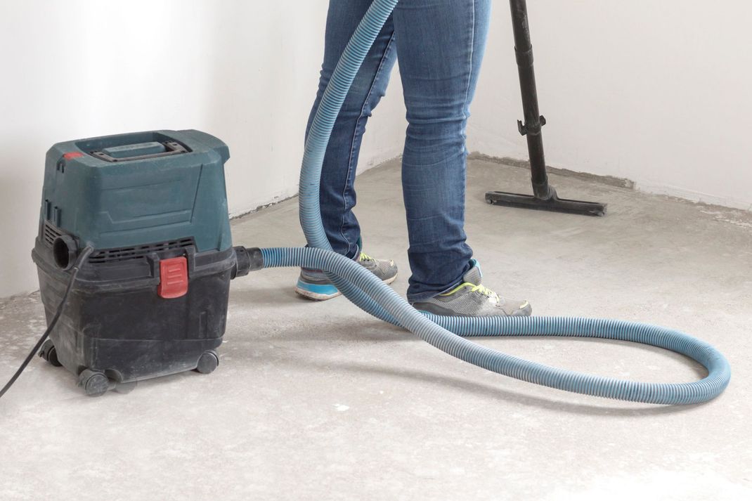 Un aspirapolvere blu con tubo flessibile su un pavimento polveroso, accanto alle gambe di una persona e al braccio di una levigatrice a muro.