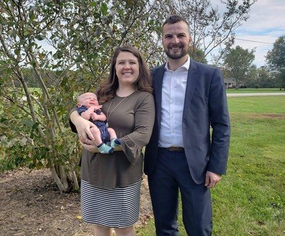 Couple with newborn baby outdoors: Woman holding baby, man in suit. Green grass, trees in background.