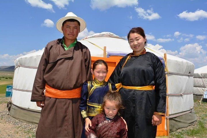 Mongolian family in traditional dress stands outside a yurt, smiling.