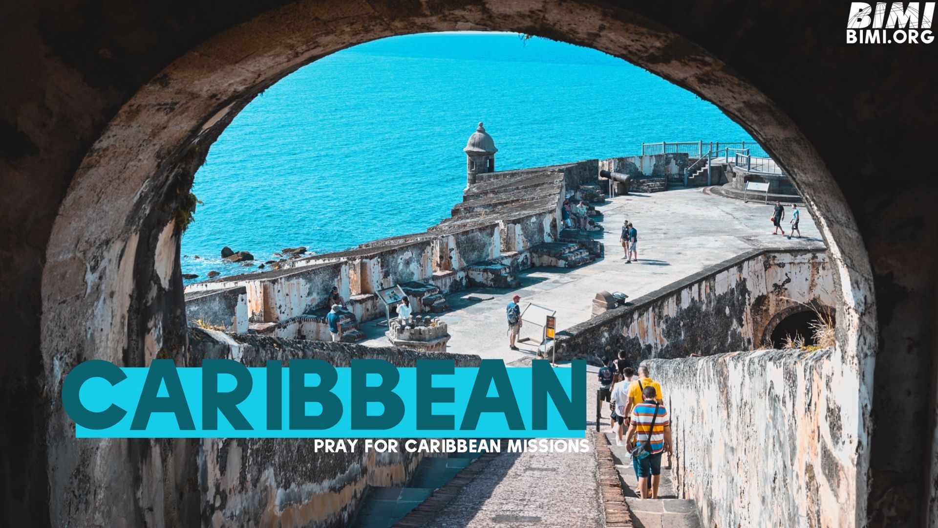 View through a stone archway of a fortress on a coast; turquoise water in the background.